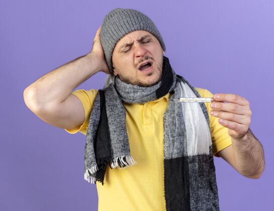 体温计戴着冬天帽子和围巾的年轻金发病患 手放在头上 看着隔离在紫色墙上的温度计冬天表情疼痛