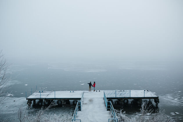 树美丽的年轻夫妇站在码头上 看着结冰的湖面天空湖家庭