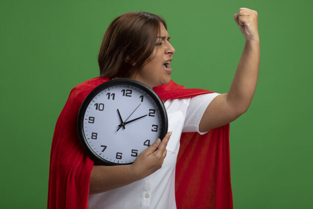 握着自信的中年女超人拿着挂钟表现出坚强的姿态孤立在绿色上时钟超级英雄手势