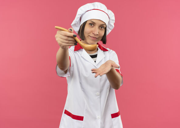 感情身着厨师制服的年轻女厨师高兴地向前伸出勺子 在粉红色的墙上显示出孤立的空手制服请人