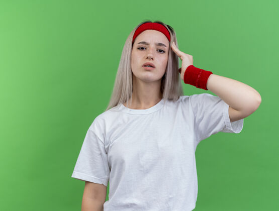 运动自信的年轻运动女性戴着背带 戴着头带和腕带 手放在隔离在绿色墙上的太阳穴上背带手女人