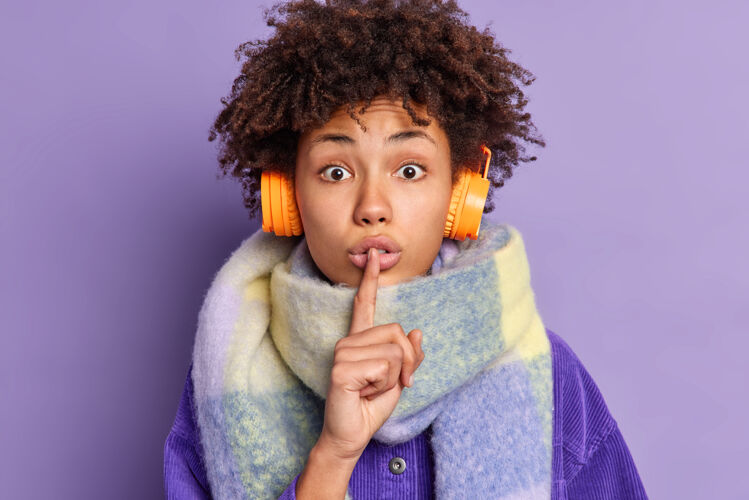 显示惊讶的非洲裔美国妇女将食指按在嘴唇上做沉默的手势告诉秘密脖子上戴着暖和的围巾通过无线耳机听音乐耳机表情人类