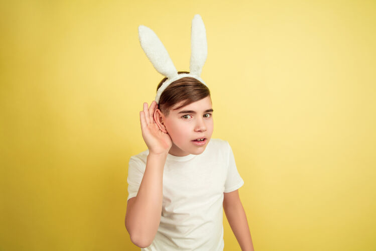 创意听秘密白人男孩作为一个复活节兔子在黄色的工作室背景复活节快乐的问候美丽的男模人类情感的概念 面部表情 节日复制空间姜搞笑男孩