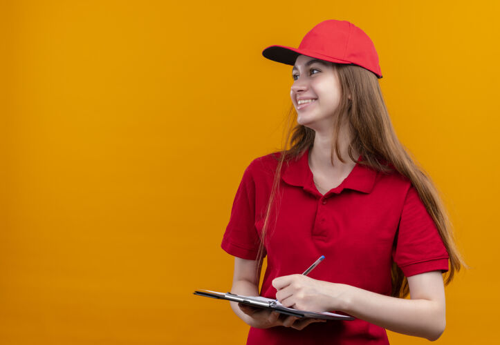 持有微笑着的年轻送货女孩穿着红色制服 拿着剪贴板和笔 看着左边孤立的橙色空间和复印空间钢笔橙色女孩