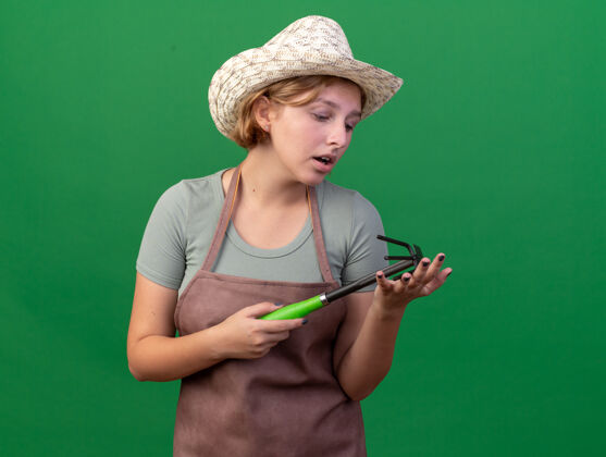 花园悲伤的年轻斯拉夫女园丁戴着园艺帽 拿着锄头看着耙子上的绿色斯拉夫女性年轻