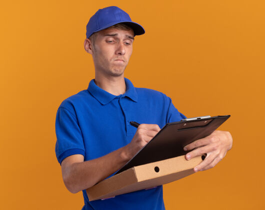 盒子自信的年轻金发送货员拿着笔写在剪贴板上的比萨饼盒上的橙色写作钢笔剪贴板
