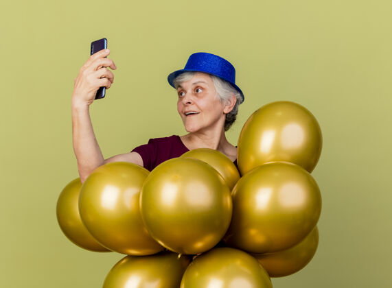举行惊讶的戴着派对帽的老妇人手持氦气球 看着隔离在橄榄绿墙上的留有复印空间的手机氦老人穿