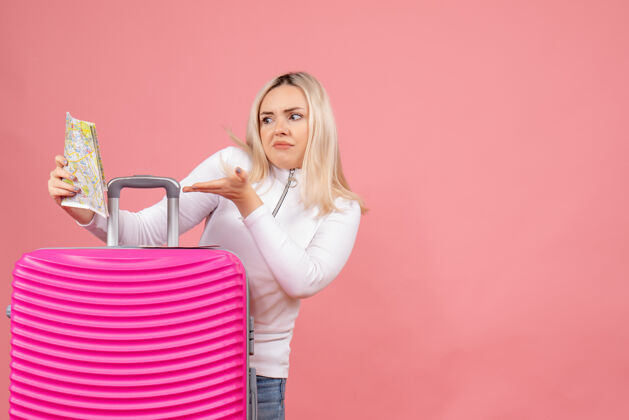 手提箱前视图困惑的年轻女士站在粉红色手提箱后面看地图微笑模特女士
