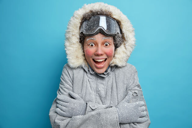 天气寒冷的冬季天气兴奋欢快的民族年轻女子 冻红了脸 在寒冷的天气里拥抱自己取暖 穿着保暖夹克和滑雪板护目镜 对一些情绪有反应雪寒假圣诞节
