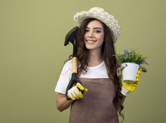 园艺微笑的年轻女园丁穿着制服 戴着园艺帽和手套 肩上拿着花盆和铁锹 隔离在橄榄绿的墙上 留着复制空间肩铁锹手套