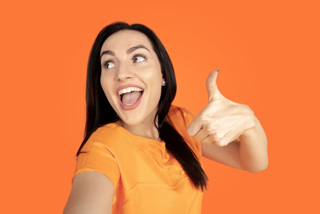 面部橙色工作室背景上的白种人年轻女子肖像穿着衬衫的漂亮黑发女性模特人类情感的概念 面部表情 销售 广告广告空间指向 展示 微笑身长衬衫黑发
