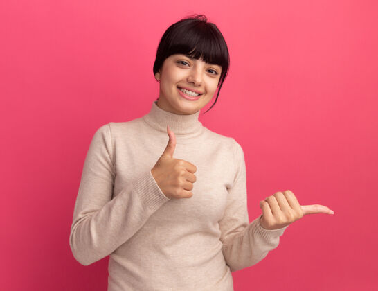 毛衣年轻的黑发白人女孩微笑着竖起大拇指 指着粉色的边黑发尖年轻