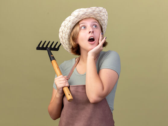 园艺兴奋的年轻斯拉夫女园丁戴着园艺帽把手放在脸上 拿着耙子看着橄榄绿的一面壁板女性帽子