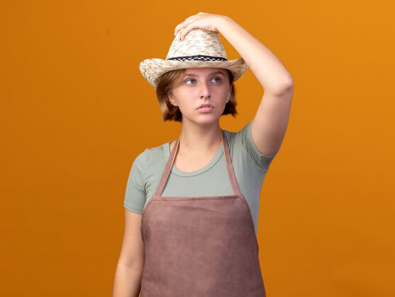 壁板自信的年轻斯拉夫女园丁戴着园艺帽把手放在帽子上看着橙色的一面花园帽子斯拉夫