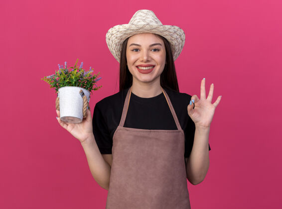 漂亮微笑着美丽的白人女园丁戴着园艺帽拿着花盆 在粉色上做着ok的手势园艺女的好的