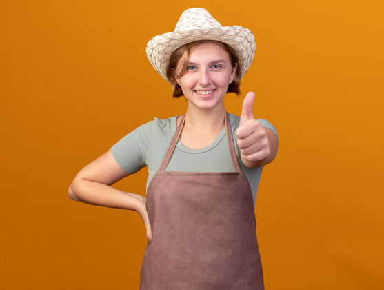 花园微笑着的年轻的斯拉夫女园丁戴着园艺帽竖起大拇指在橘子上帽子年轻的斯拉夫人