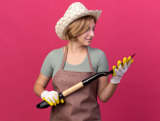黑桃高兴的年轻斯拉夫女园丁戴着园艺帽和手套拿着和看着粉红色的铲子园艺拜托帽子