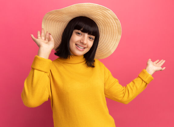 帽子高兴的年轻黑发白人女孩戴着沙滩帽站在粉红色上举手黑发人站起来举起手来