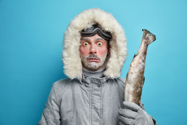 胡德受惊情绪激动的男子满脸通红 身上覆盖着白霜 冬季远征时去钓鱼 手里拿着大鱼 穿着夹克 戴着滑雪镜极端冰冻钓鱼