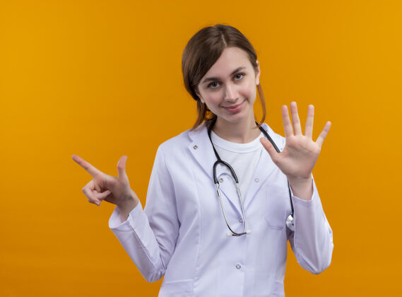 长袍高兴的年轻女医生穿着医用长袍和听诊器指着左边 并显示在孤立的橙色空间5穿请听诊器