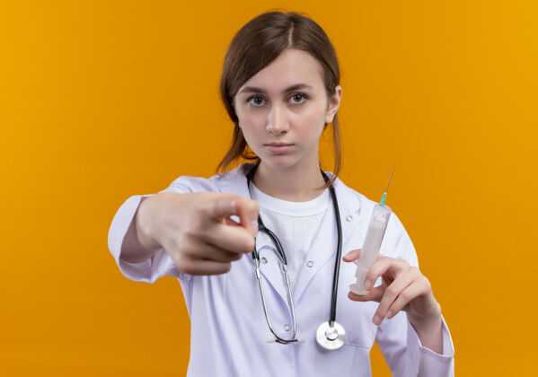 医生面容严肃的年轻女医生穿着医用长袍和听诊器拿着注射器 指着孤立的橙色空间听诊器指着拿着