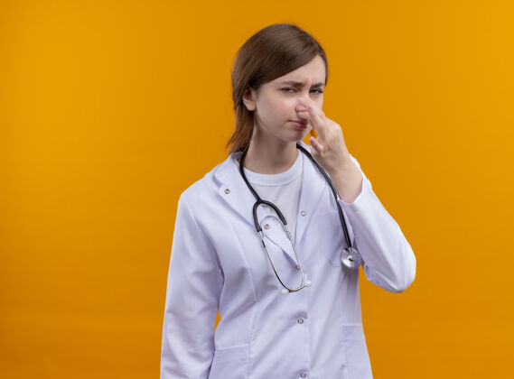 太空恼怒的年轻女医生穿着医用长袍和听诊器拿着鼻子在孤立的橙色空间与复印空间鼻子女性拿着