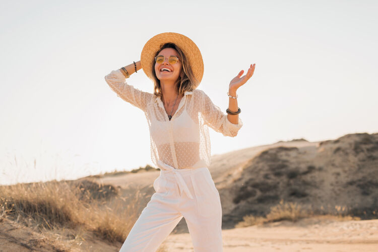 旅行时尚快乐的美丽微笑的女人在沙漠里摆姿势 穿着白色的衣服 戴着草帽和太阳镜在夕阳下太阳镜帽子时尚