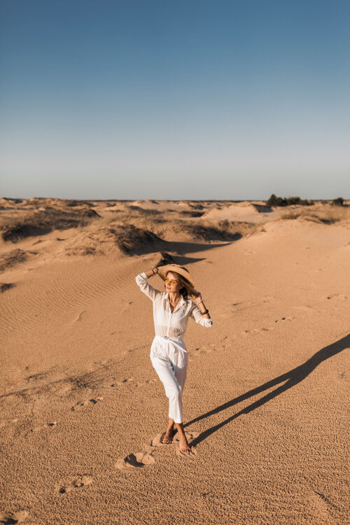 旅游日落时分 穿着白色裤子和衬衫 头戴草帽 走在沙漠沙滩上的时髦美女休闲夏天人