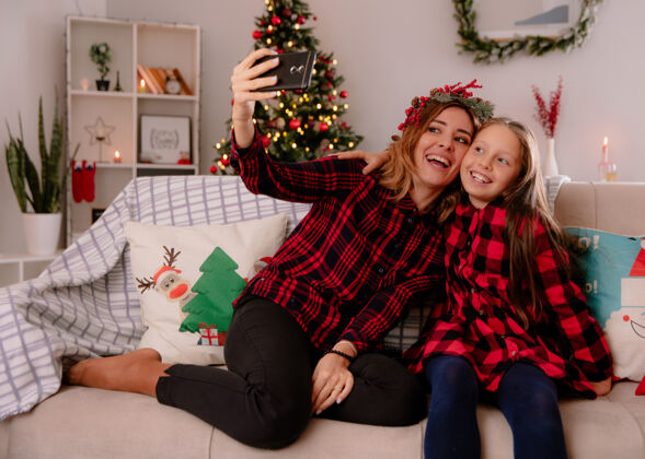 圣诞快乐带着冬青花环的笑妈妈和女儿看着手机坐在沙发上自拍 在家里享受圣诞节时光花圈时间享受