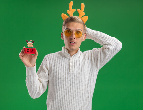 驯鹿令人印象深刻的年轻帅哥戴着驯鹿鹿角头带戴着眼镜拿着圣诞树玩具和日期看着相机保持手在头后面隔离在绿色背景上绿色年轻人头带