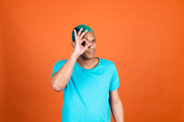 表情黑色非洲男人在橙色的墙上随意的蓝色头发显示ok手势遮住一只眼睛情绪时髦洞