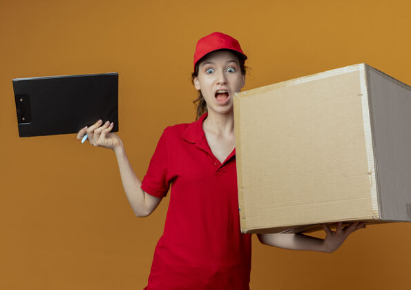 红色令人印象深刻的年轻漂亮的送货女孩在红色制服和帽子拿着钢笔和纸板盒向下看橙色背景孤立剪贴板橙色钢笔女孩