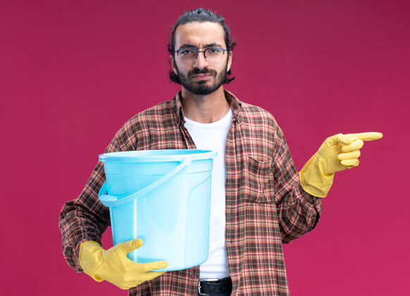 姿势年轻帅气的清洁工穿着t恤 戴着手套 手里拿着水桶 指着隔离在粉红色墙上的一边点站感情