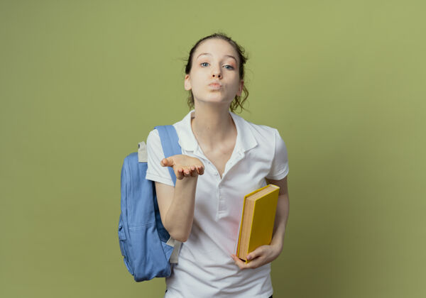背景自信的年轻漂亮的女学生 背着书包 拿着书 对着隔离在绿色背景下的相机吹吻 还有复印空间漂亮发送吹