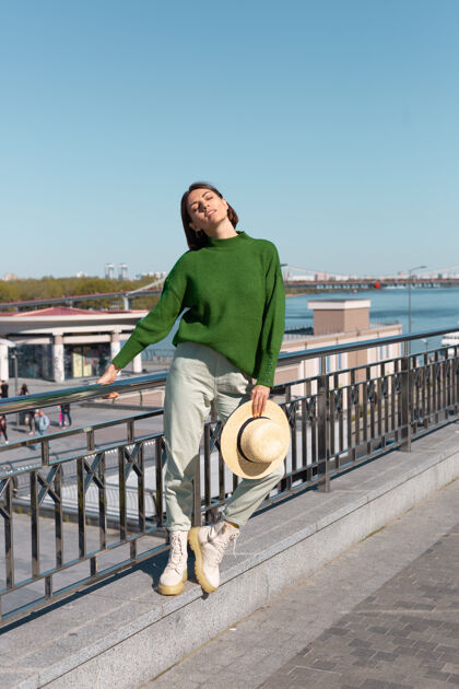 自由穿着绿色休闲毛衣 戴着帽子的时尚女士在户外的桥上欣赏河景 享受夏日阳光明媚的日子人女士姿势