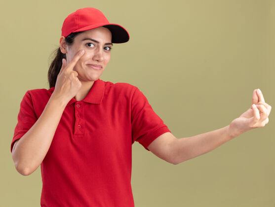 提示令人高兴的年轻送货女孩穿着制服 戴着帽子 手指放在眼睛上 在橄榄绿的墙上显示着孤立的小费手势帽子姿势手指