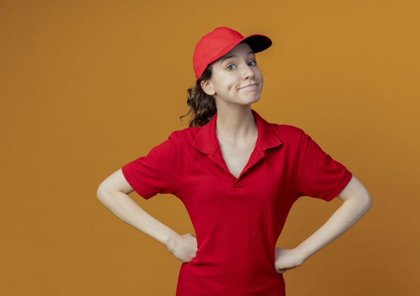 橙色令人印象深刻的年轻漂亮的送货女孩在红色制服和帽子把手放在腰上看着相机孤立的橙色背景与复制空间帽子印象红色