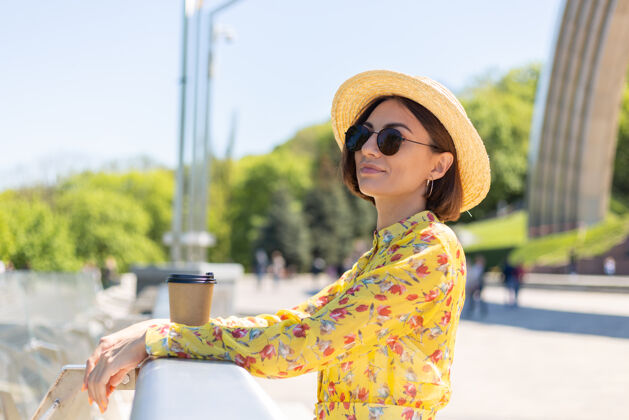 城市景观穿着黄色夏装 戴着黄色帽子 喝着咖啡 享受阳光的女人的户外肖像 站在桥上 城市的景色令人惊叹地标姿势城镇