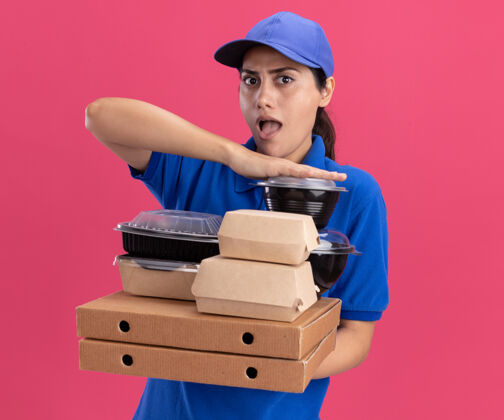 送货惊讶的年轻送货女孩穿着制服 戴着帽子 把食物容器放在隔离在粉红色墙上的比萨饼盒上姿势食物披萨