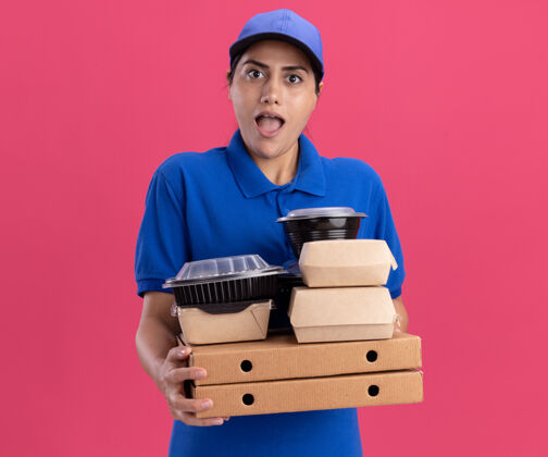 人惊讶的年轻送货女孩穿着制服 戴着帽子 把食物容器放在隔离在粉红色墙上的比萨饼盒上人年轻人惊喜