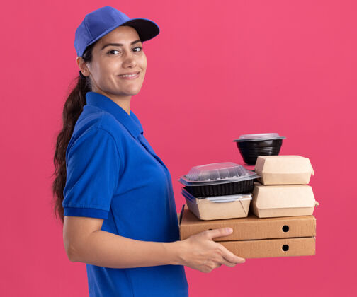 衣服微笑着站在侧视图年轻的送货女孩穿着制服 戴着帽子拿着食物容器放在比萨饼盒上 隔离在粉红色的墙上人年轻人盒子