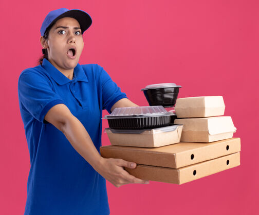 姿势被吓坏了的年轻送货女孩穿着制服 戴着帽子 拿着比萨饼盒上的食物容器 隔离在粉红色的墙上年轻人帽子盒子