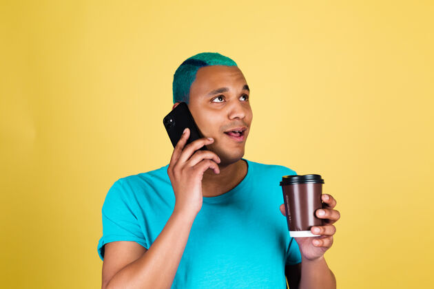 西班牙人非洲黑人男人在随意的黄墙上蓝色的头发上享受着一杯咖啡 积极快乐的情绪在电话里谈笑风生男人茶高兴起来