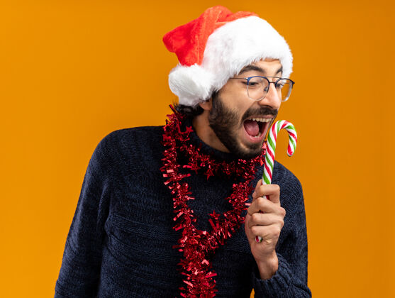 年轻人兴奋的年轻帅哥戴着圣诞帽 脖子上戴着花环 手里拿着橙色背景的圣诞糖果脖子糖果兴奋