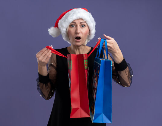 圣诞快乐印象深刻的中年金发女人戴着圣诞帽拿着圣诞礼品袋打开一个看着相机孤立的紫色背景女人包打开