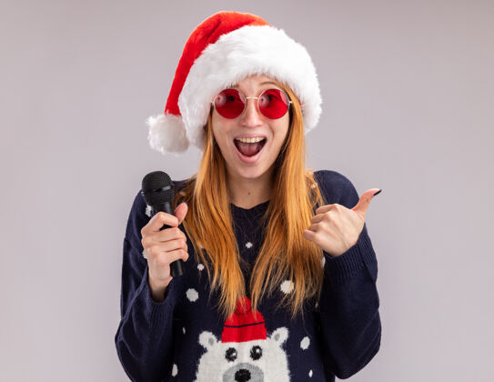 帽子兴奋的年轻漂亮女孩戴着圣诞帽戴着眼镜拿着麦克风在白色背景上孤立地竖起大拇指圣诞快乐显示年轻
