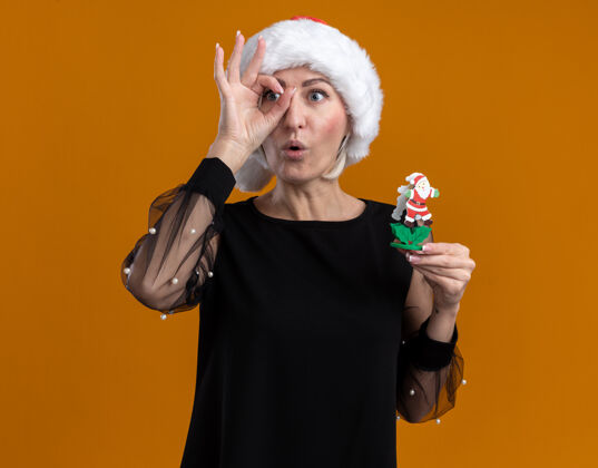 圣诞节令人印象深刻的中年金发女人戴着圣诞帽看着一边拿着圣诞老人玩具做样子手势孤立在橙色背景上帽子圣诞老人壁板