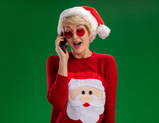 女人令人印象深刻的年轻金发女人戴着圣诞帽和圣诞老人的圣诞毛衣戴着眼镜讲电话看着绿色背景下孤立空间年轻帽子