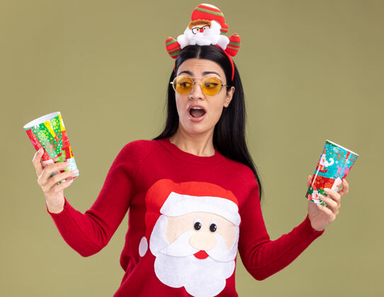 塑料印象深刻的年轻白人女孩戴着圣诞老人的头带和毛衣 戴着眼镜 举着塑料圣诞杯 看着其中一个孤立的橄榄绿背景圣诞快乐年轻一个