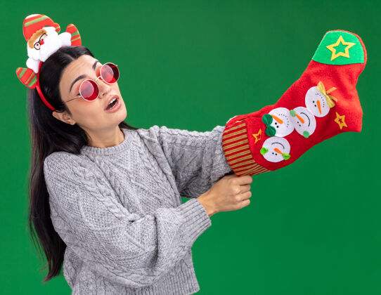 里面好奇的白人女孩戴着圣诞老人的头带 戴着眼镜 手里拿着圣诞长袜 看着它 把手放在里面 在绿色的背景上与世隔绝年轻圣诞老人眼镜